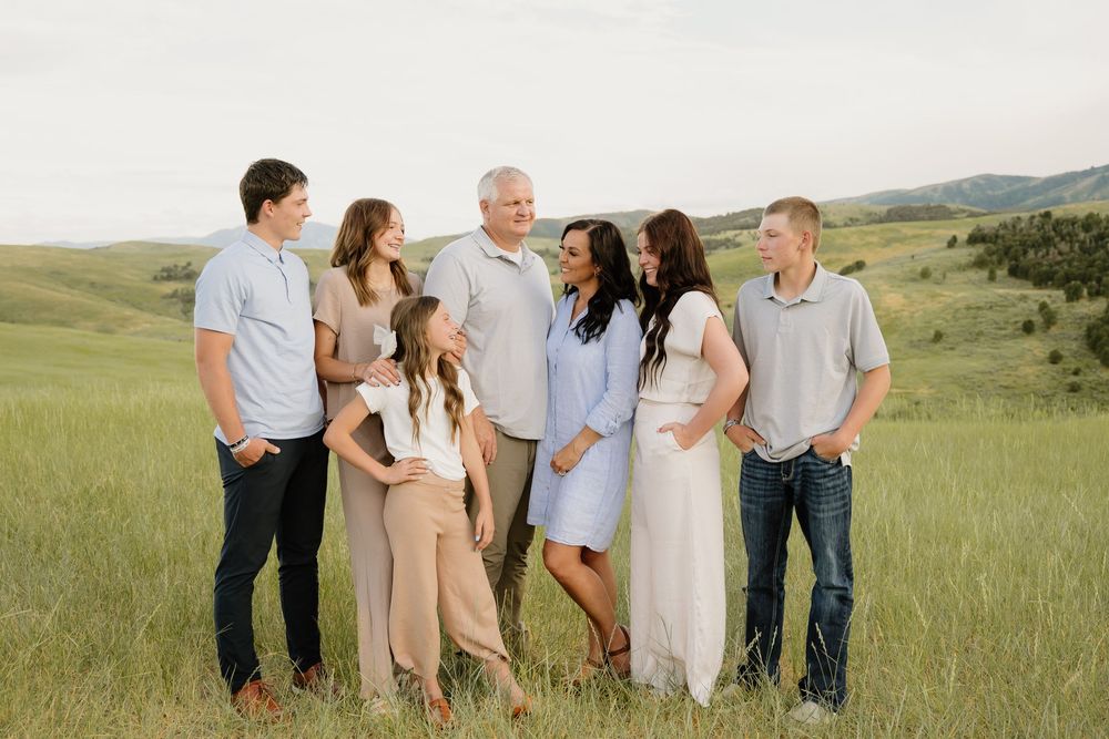 Group wearing coordinated pastel outfits stands in scenic green rolling hills during sunset for family photos.