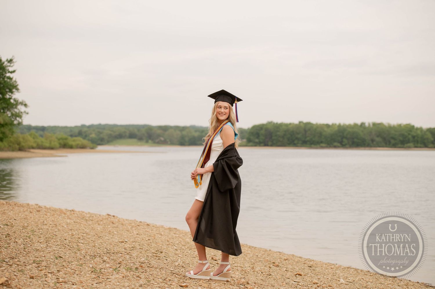 A graduate in black cap and gown stands barefoot on a sandy beach shore beside a serene lake.