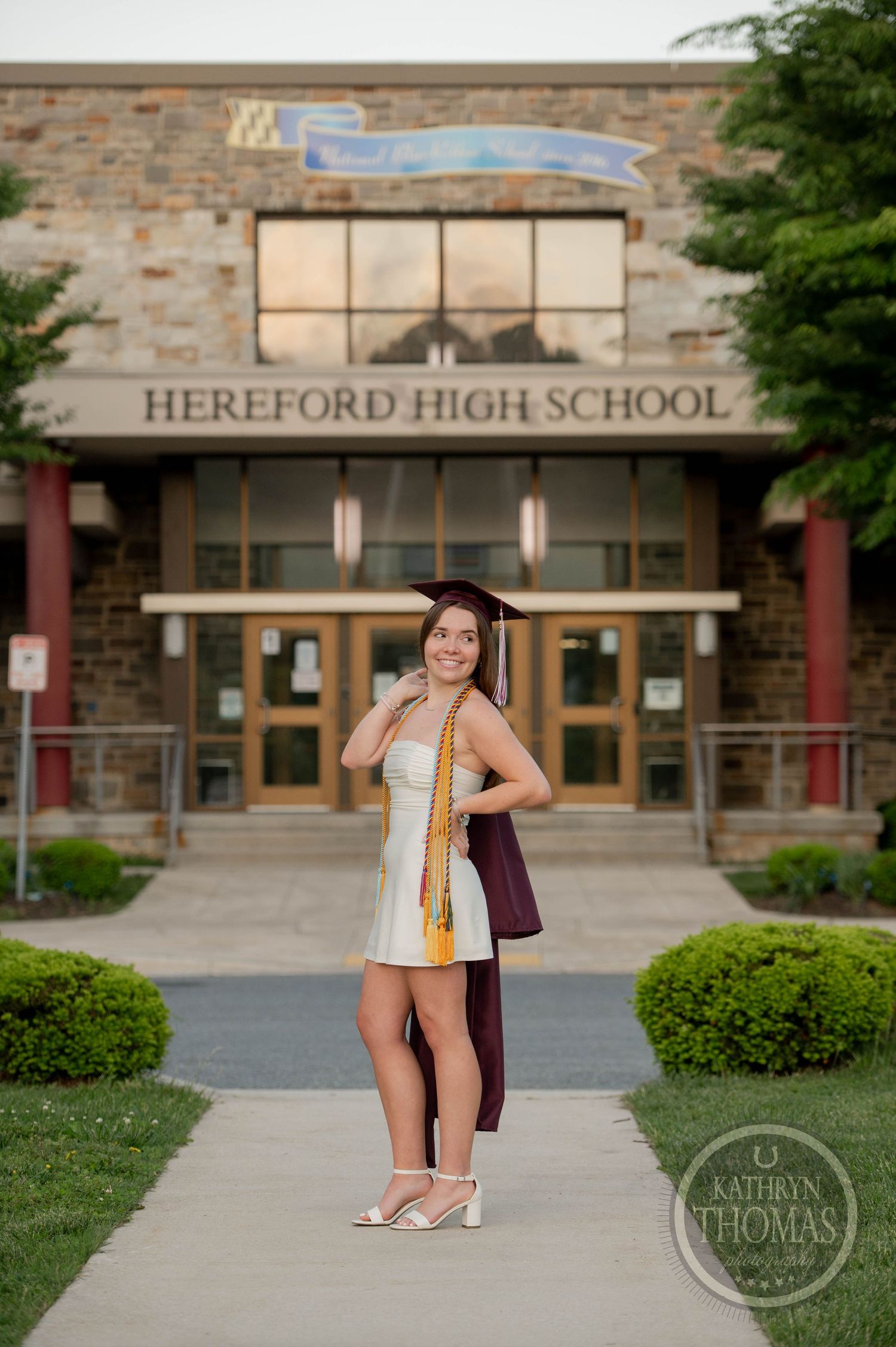 A graduating student in a white dress and stole poses in front of Hereford High School entrance on graduation day.
