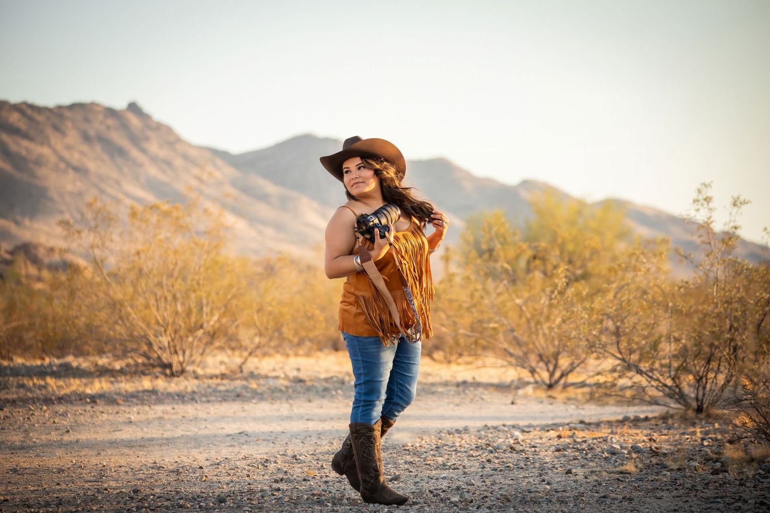 A western-style figure in denim and boots walks through a desert landscape with mountains in the background at golden hour.