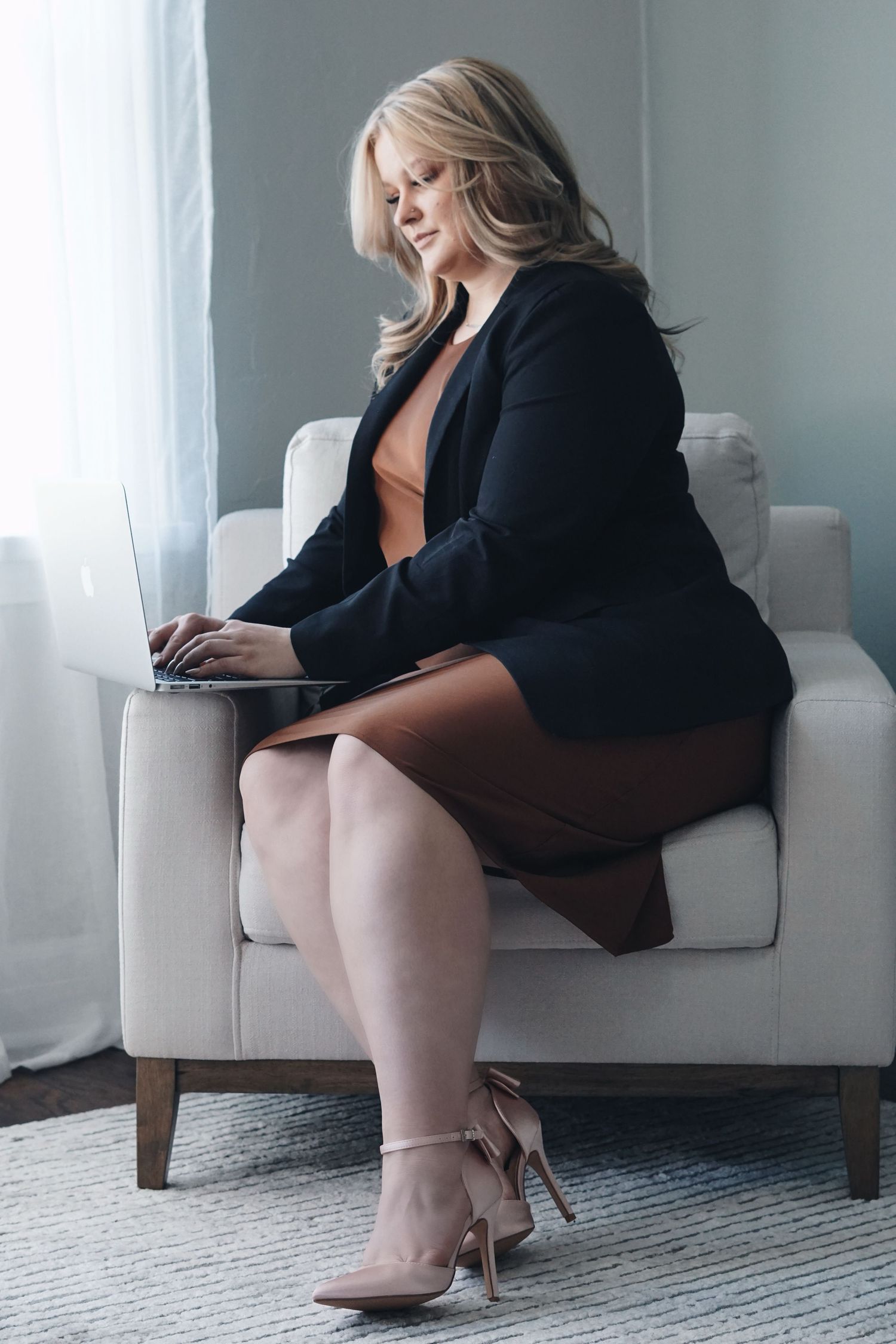 Soft lit image of a person working on laptop while seated in white armchair, wearing black blazer and beige stilettos.
