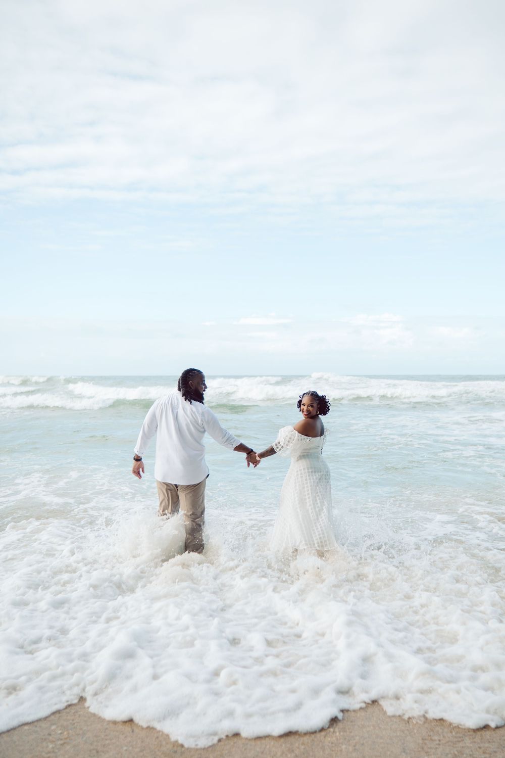 A couple in white attire holding hands while standing in the ocean waves at the beach during a romantic photo session.
