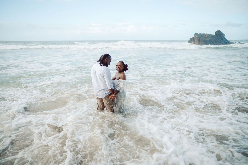 A couple in wedding attire stands in the ocean waves at the shore with a rocky island visible in the background.