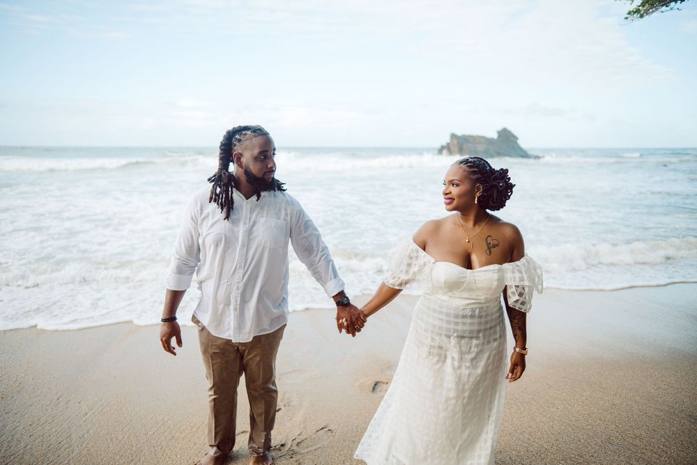 Couple walking hand in hand along a tropical beach in white wedding attire as ocean waves crash on the shore.