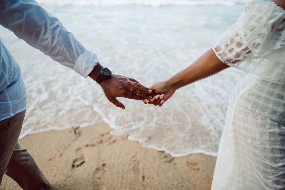 Two people holding hands while standing on a sandy beach with ocean waves gently rolling in the background.