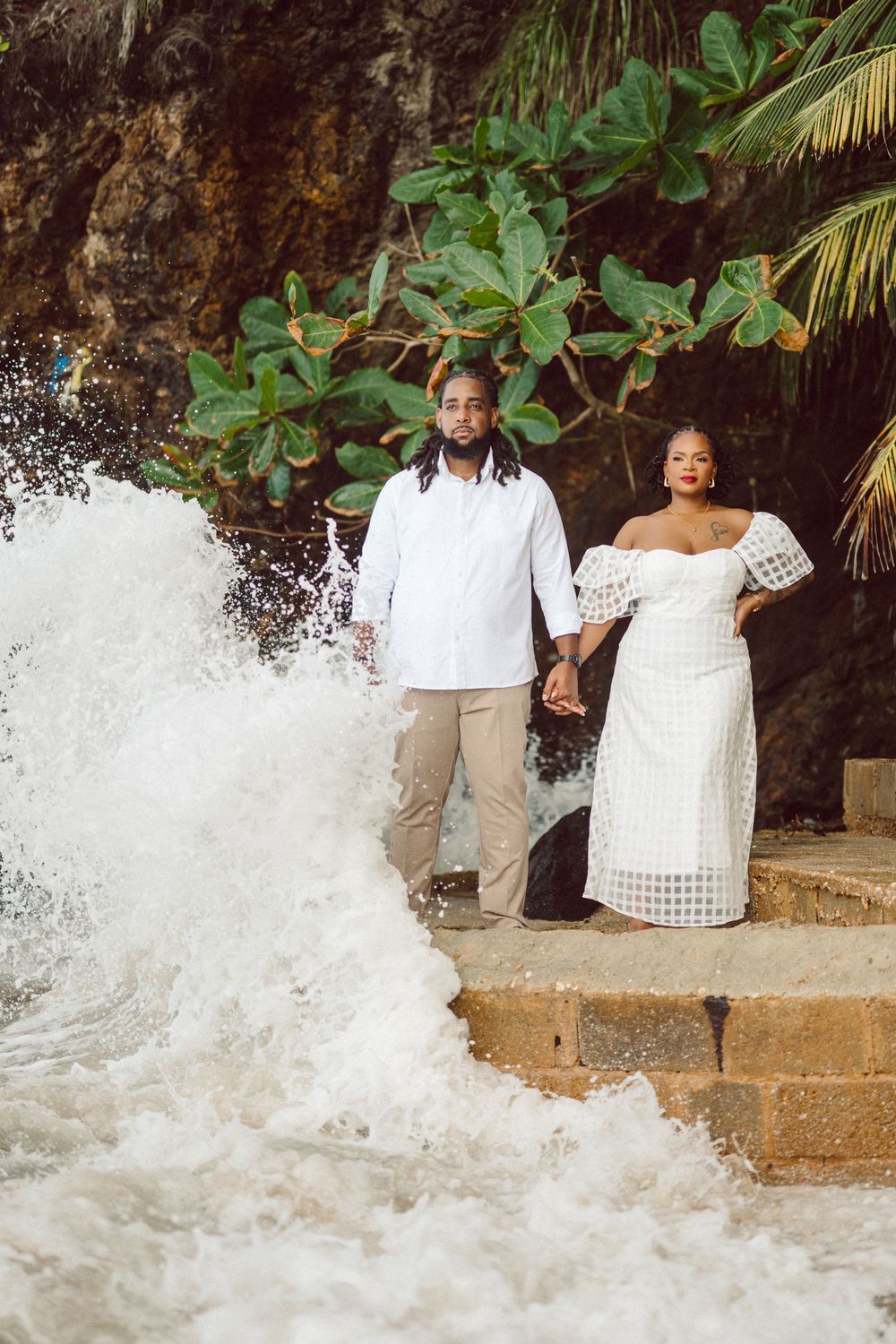 A couple stands on stone steps by crashing ocean waves against a tropical backdrop with palm trees and greenery.