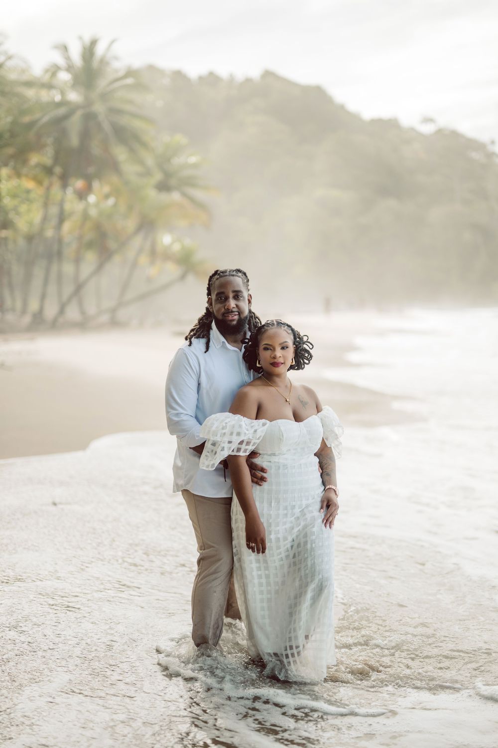 Two people embrace on a misty tropical beach with palm trees and mountains in the background during sunset.