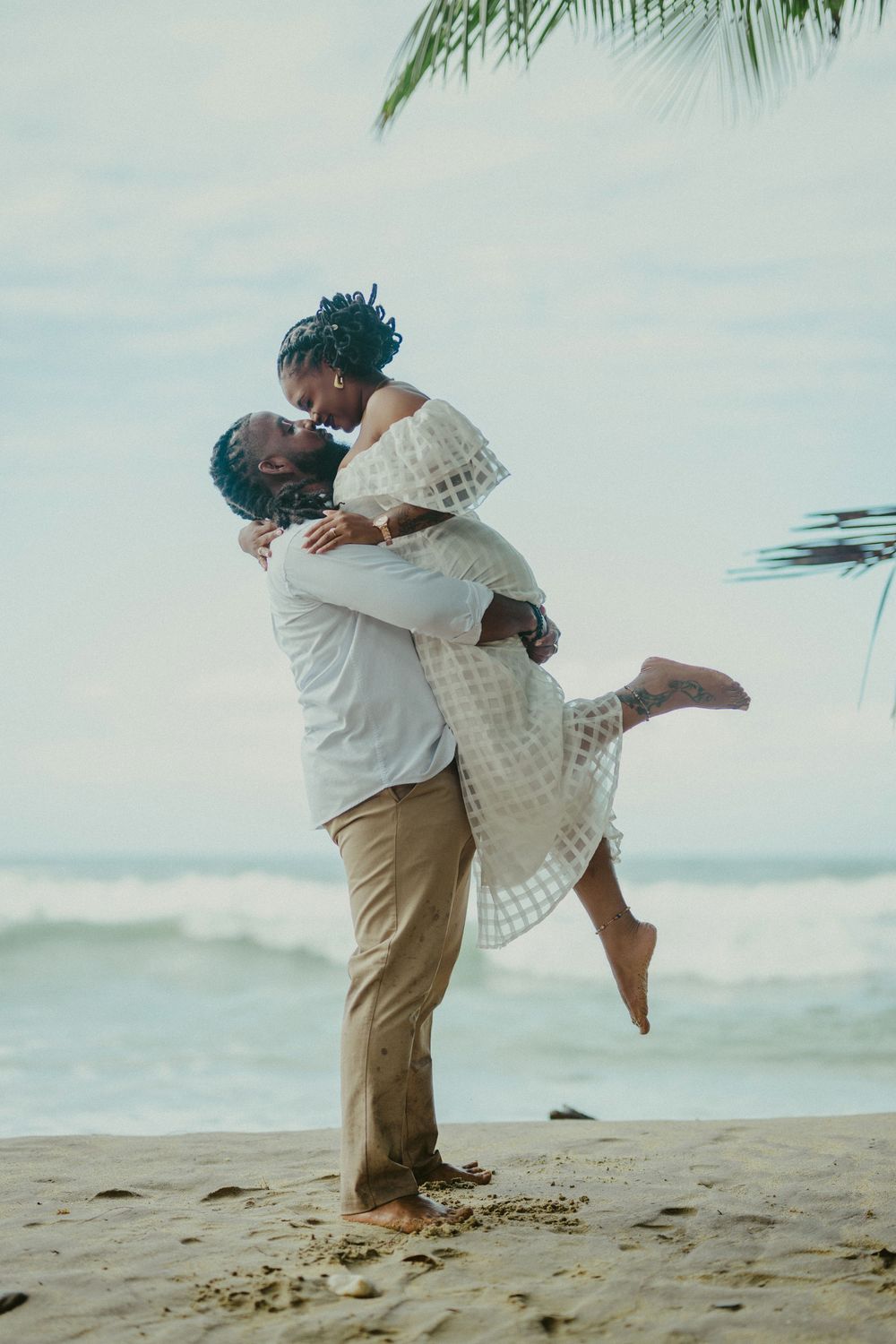 Two people in white and beige embracing romantically on a tropical beach with palm trees in the background.