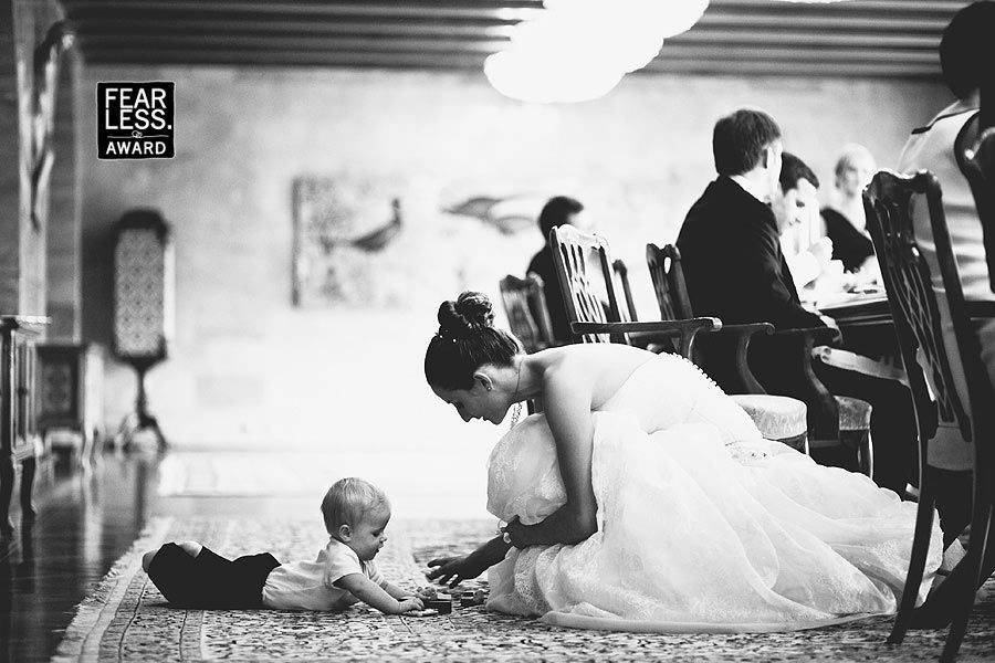 A bride in a white wedding dress bends down to interact with a baby lying on an ornate carpet during a formal event.
