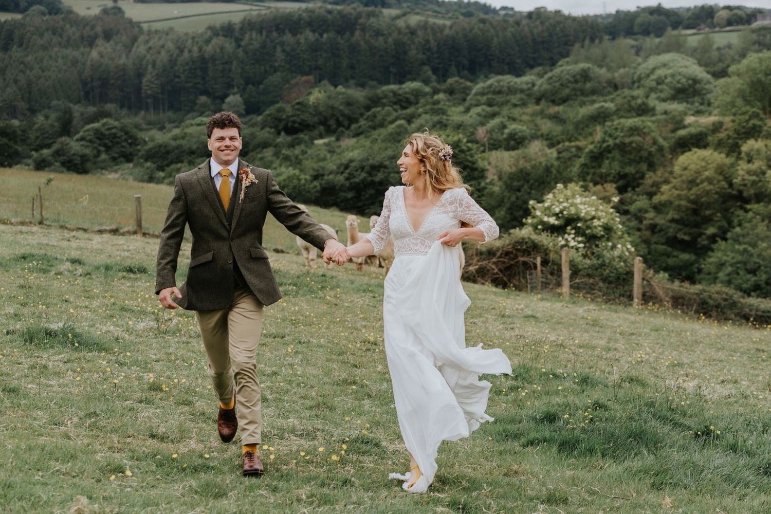 A couple runs joyfully through a lush green hillside meadow, with the bride in flowing white dress and groom in dark suit.