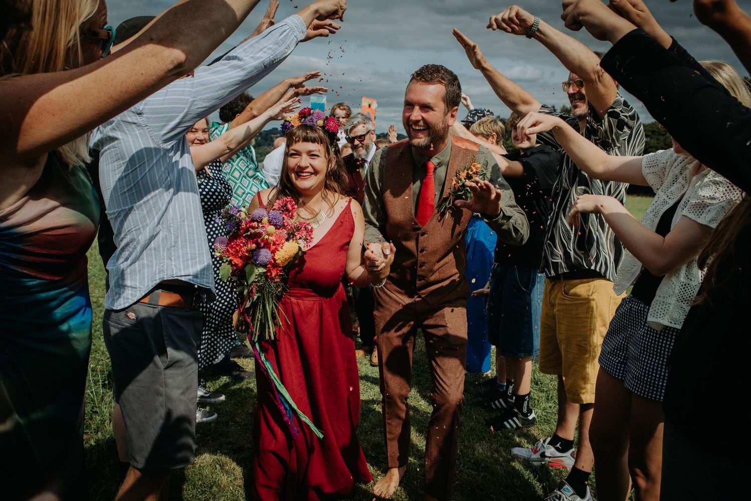 Wedding guests cheer and celebrate at an outdoor celebration as red-dressed attendees pass beneath an arch of raised hands.
