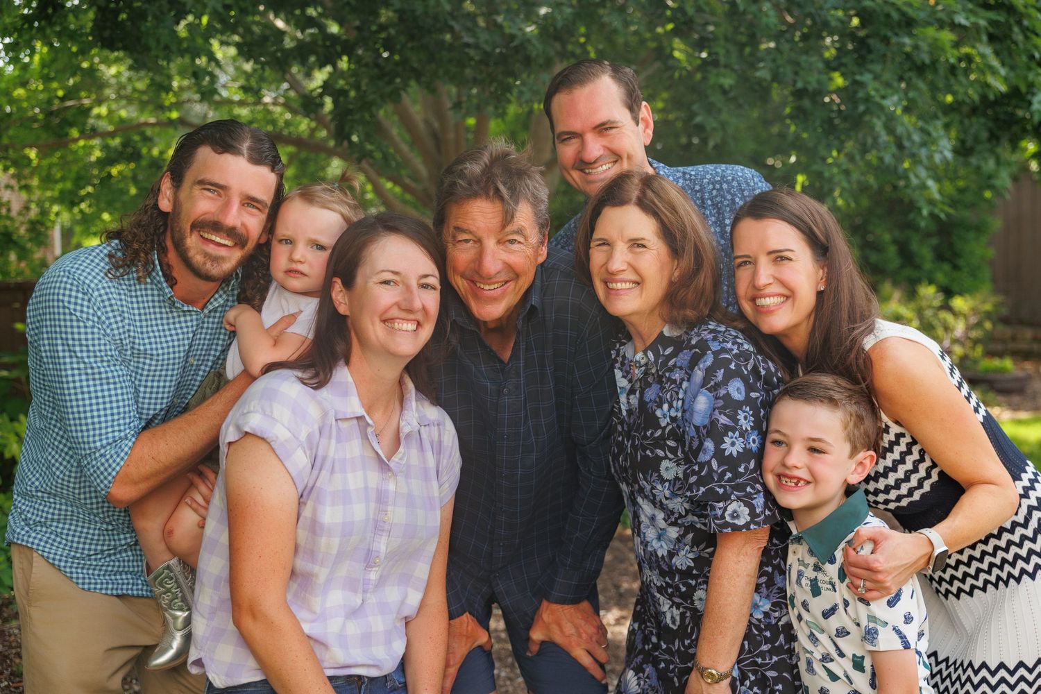 A group of happy people in casual attire gather together outdoors for a cheerful photo with green foliage in the background.