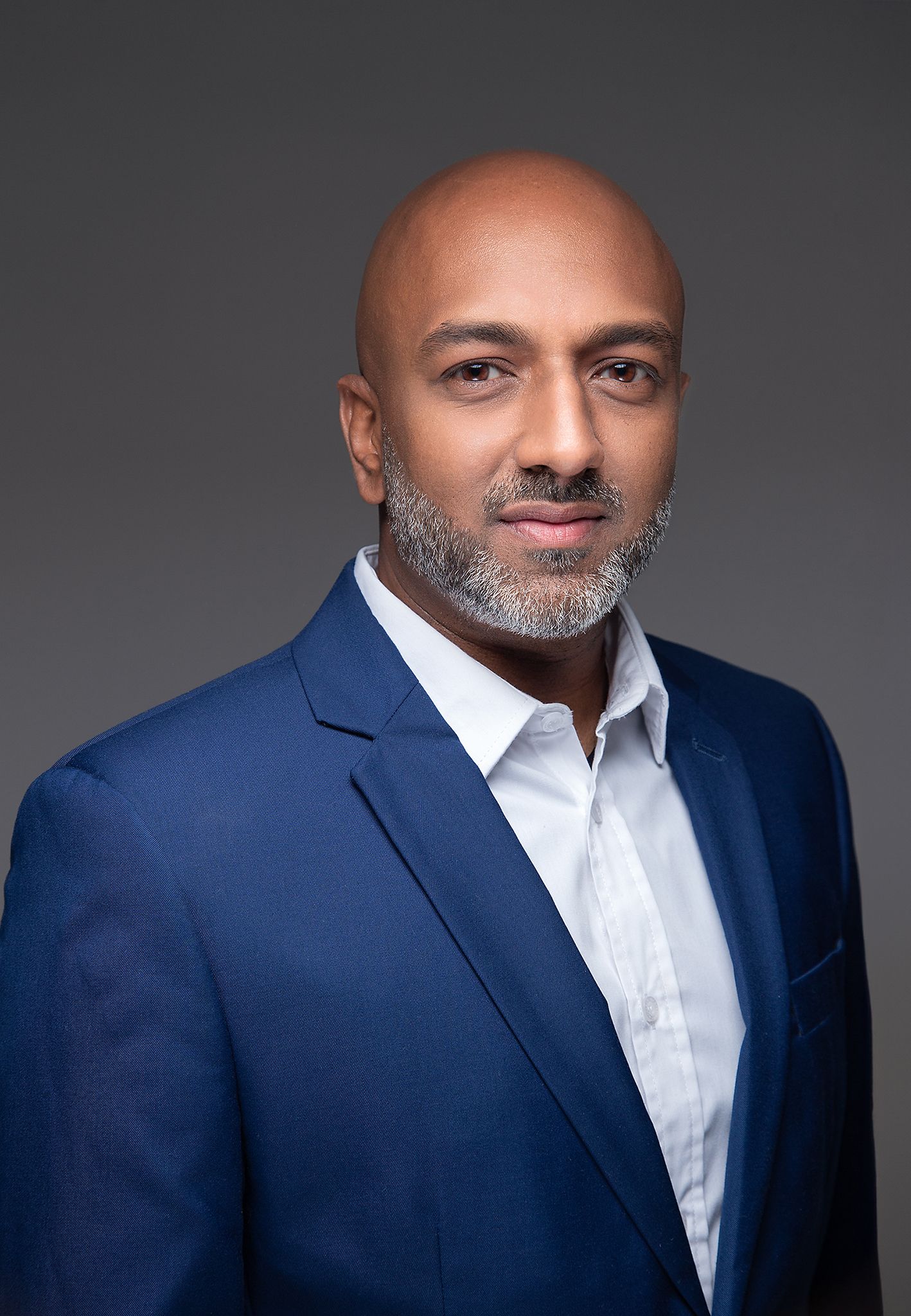 Professional portrait of someone in an all-black business suit and blazer against a gray studio background.