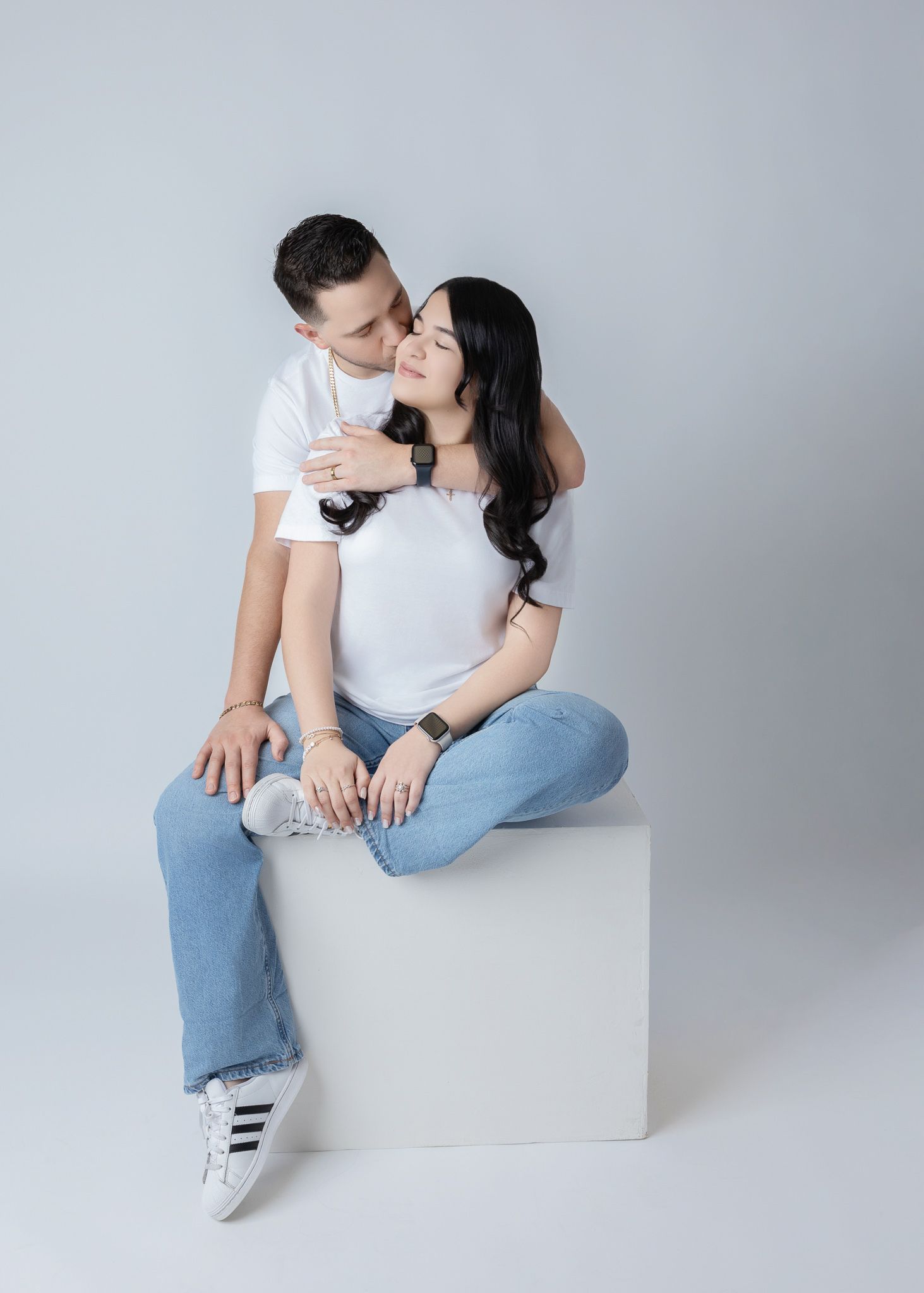 A couple in white shirts and gray pants pose playfully together in a bright minimalist studio setting.