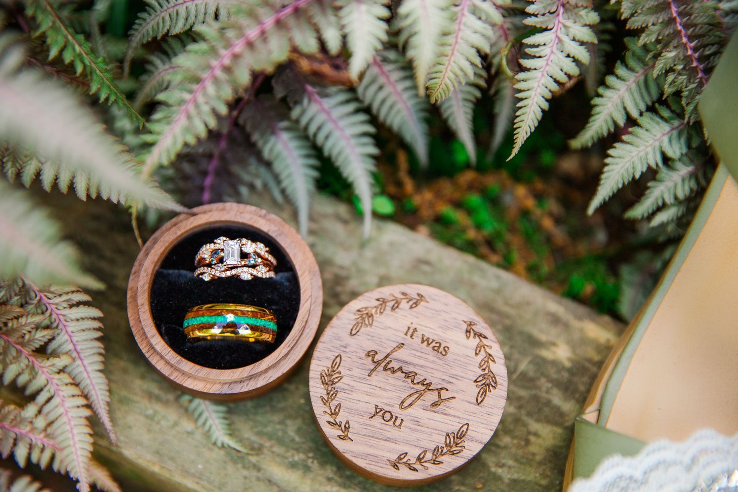 Wedding rings displayed in a wooden ring box surrounded by fern leaves on a stone surface.