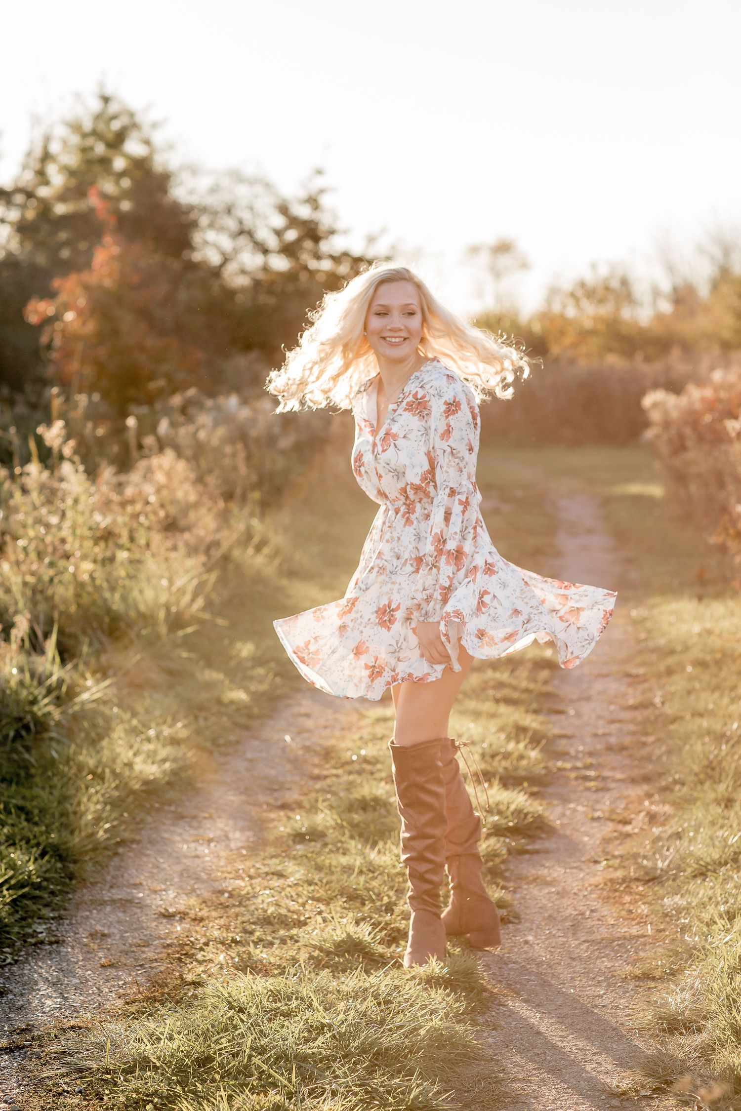 Blonde woman in floral dress and brown boots twirling on a sunlit country path in autumn.