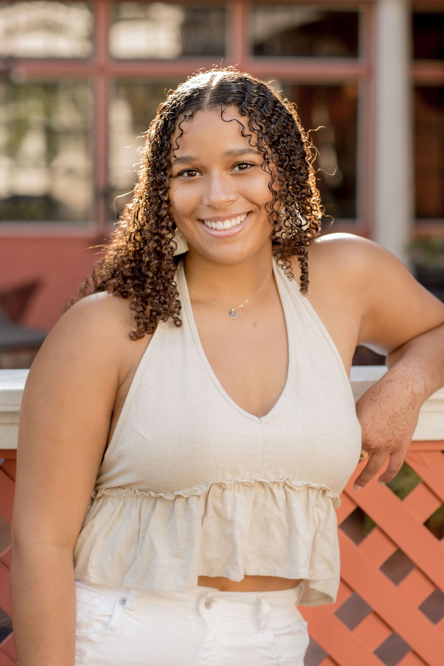 Smiling young woman with curly hair wearing a beige halter top leaning on a red fence outdoors.
