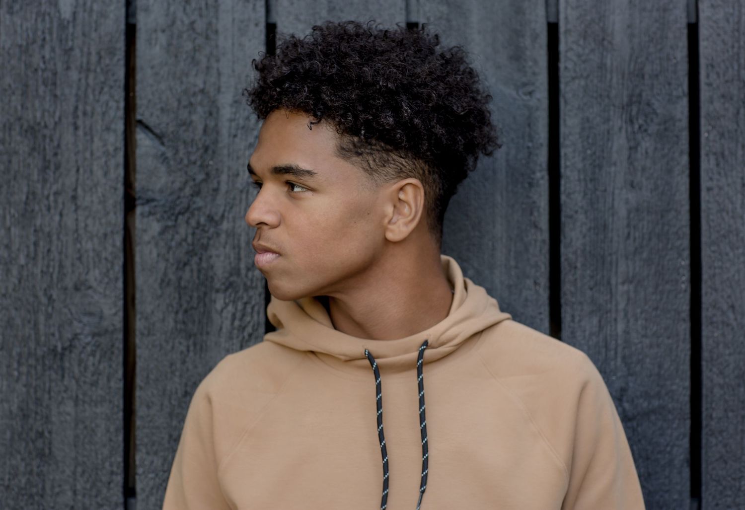 Young man in beige hoodie looking sideways against dark wooden fence background.