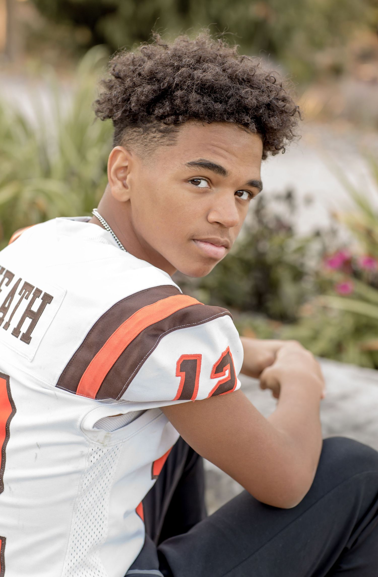 Teen boy in football jersey sitting outdoors with confident pose among garden greenery.