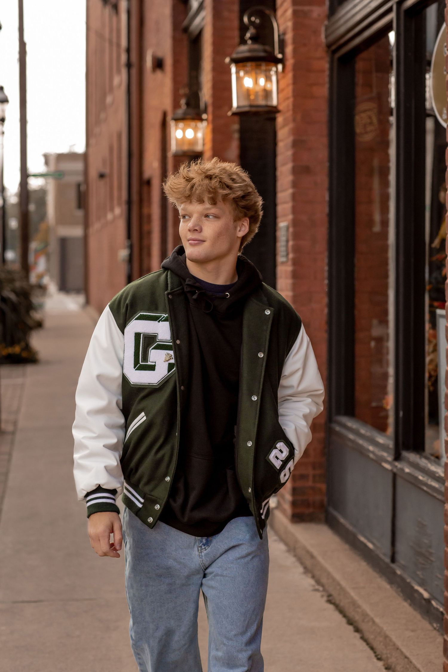 Teen boy in green and white varsity jacket walking on a brick sidewalk in an urban setting at dusk.