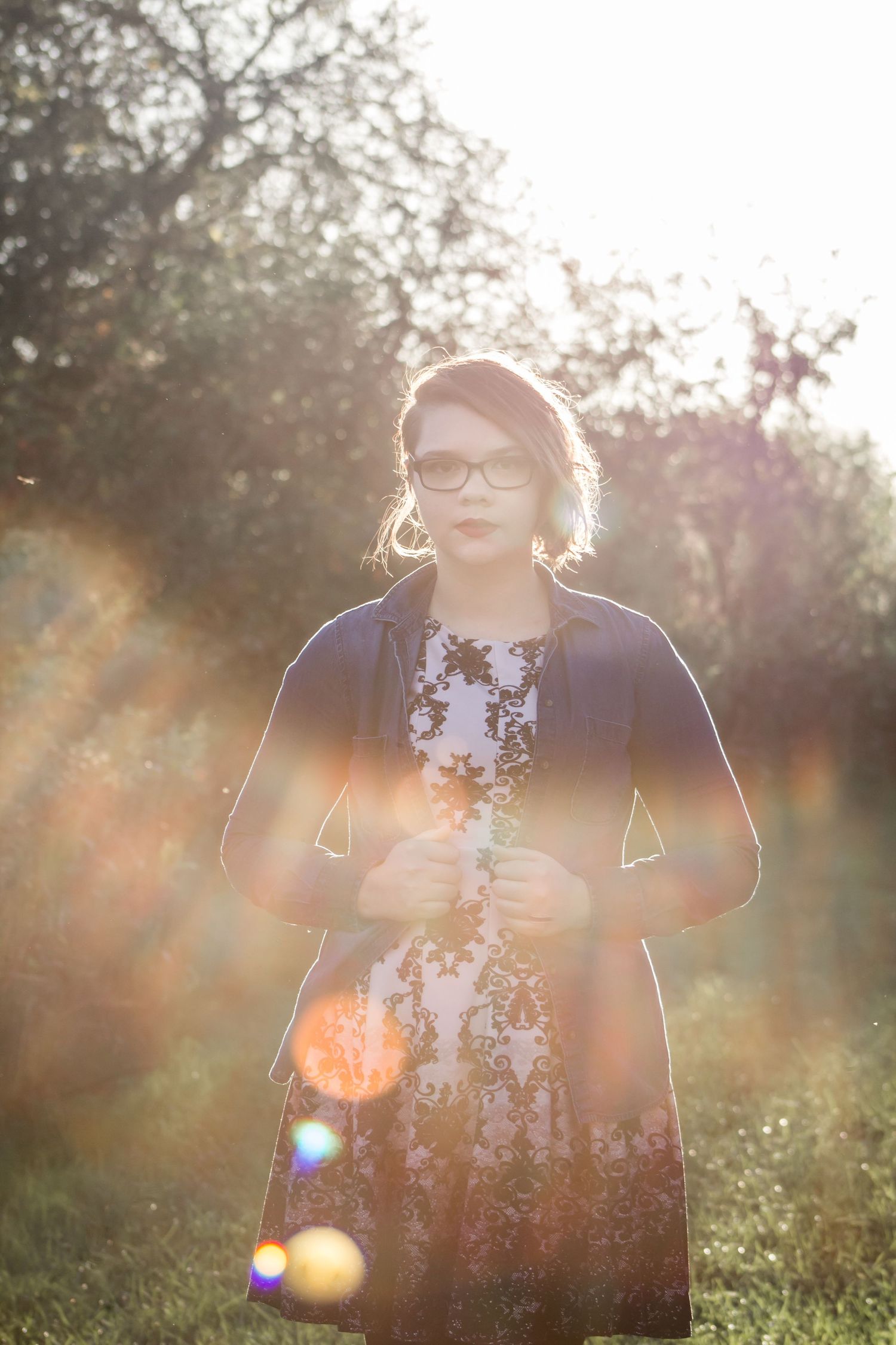 A woman in glasses and a floral dress stands in a sunlit garden with beautiful lens flare bokeh effect.