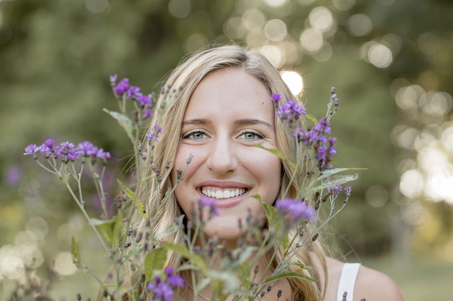 A smiling blonde woman poses with purple wildflowers in a sunny outdoor portrait session.