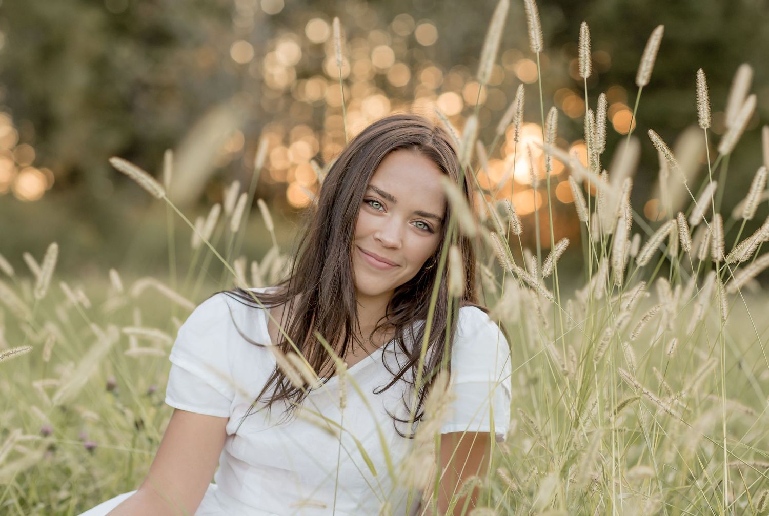 Young brunette woman sitting in tall grass field smiling softly during golden hour sunset.