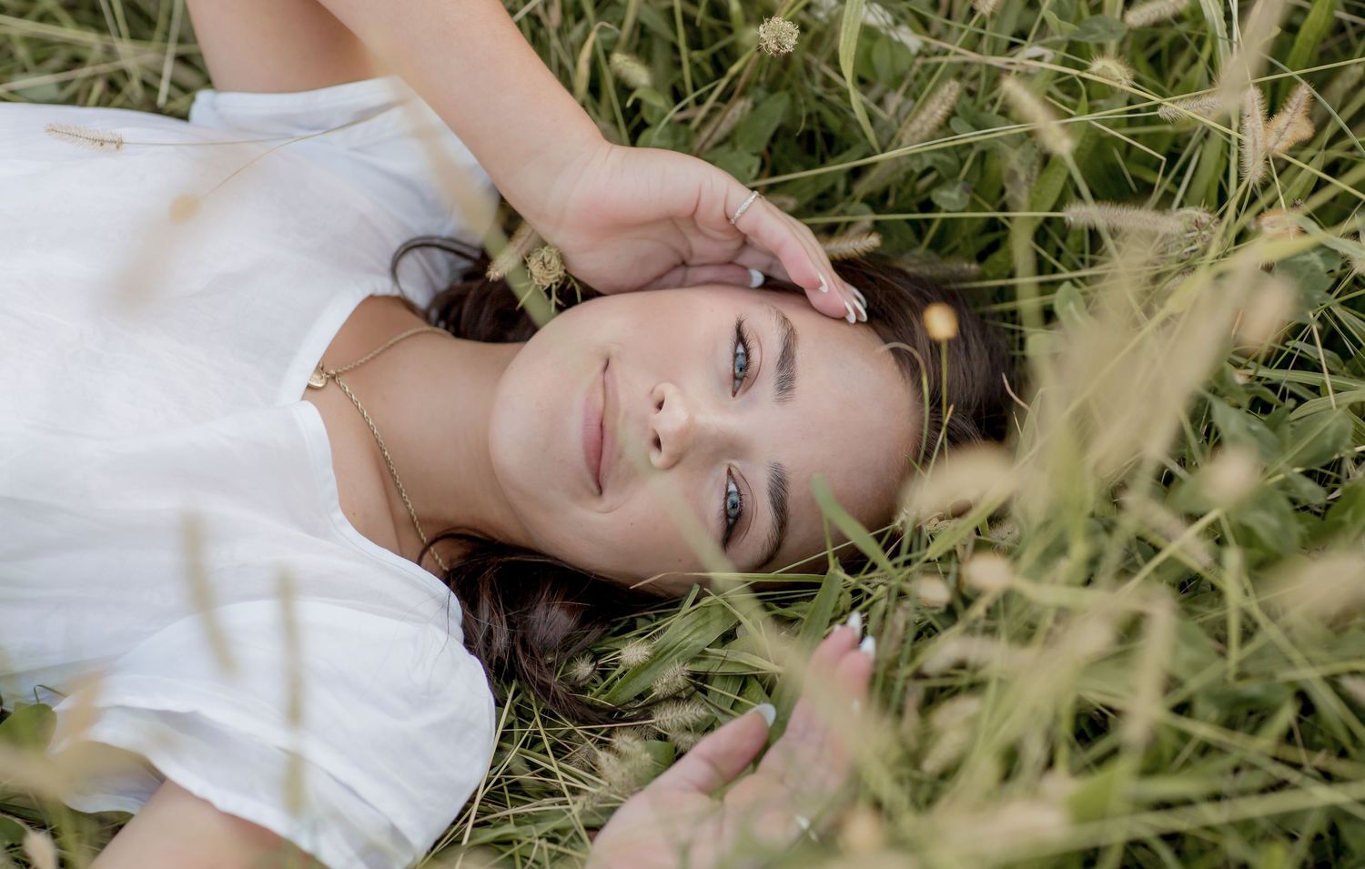 Young woman in white dress lying in tall grass field smiling up at camera.