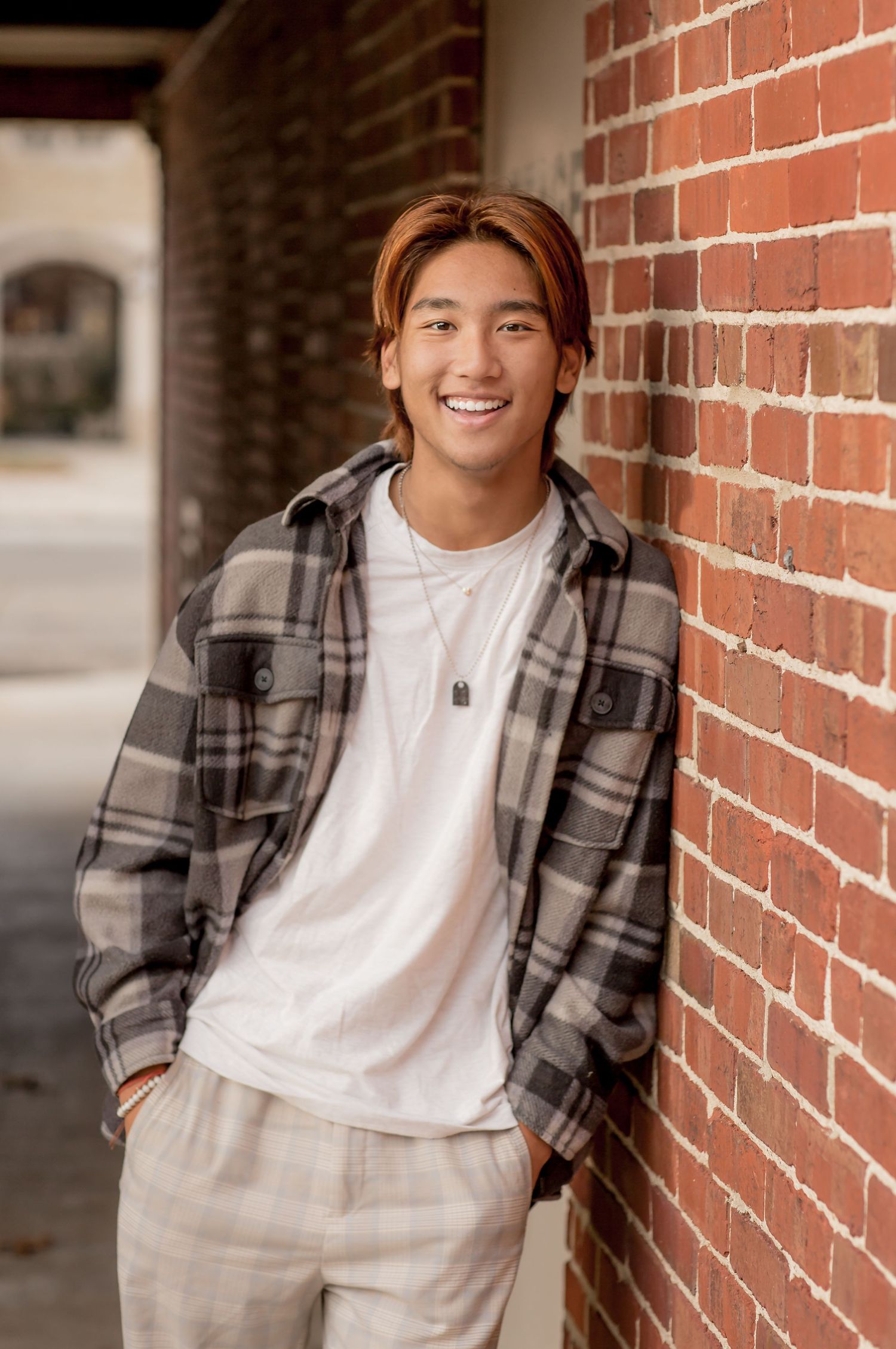 Smiling young man with auburn hair leaning against a brick wall wearing a plaid flannel shirt and white tee.