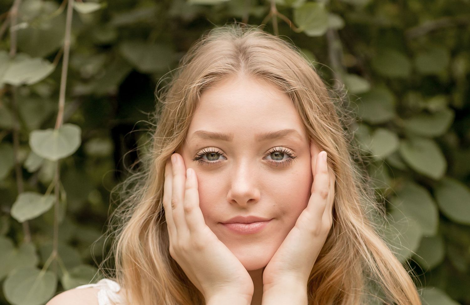 Young blonde girl with blue eyes resting her face in her hands in front of lush green foliage.