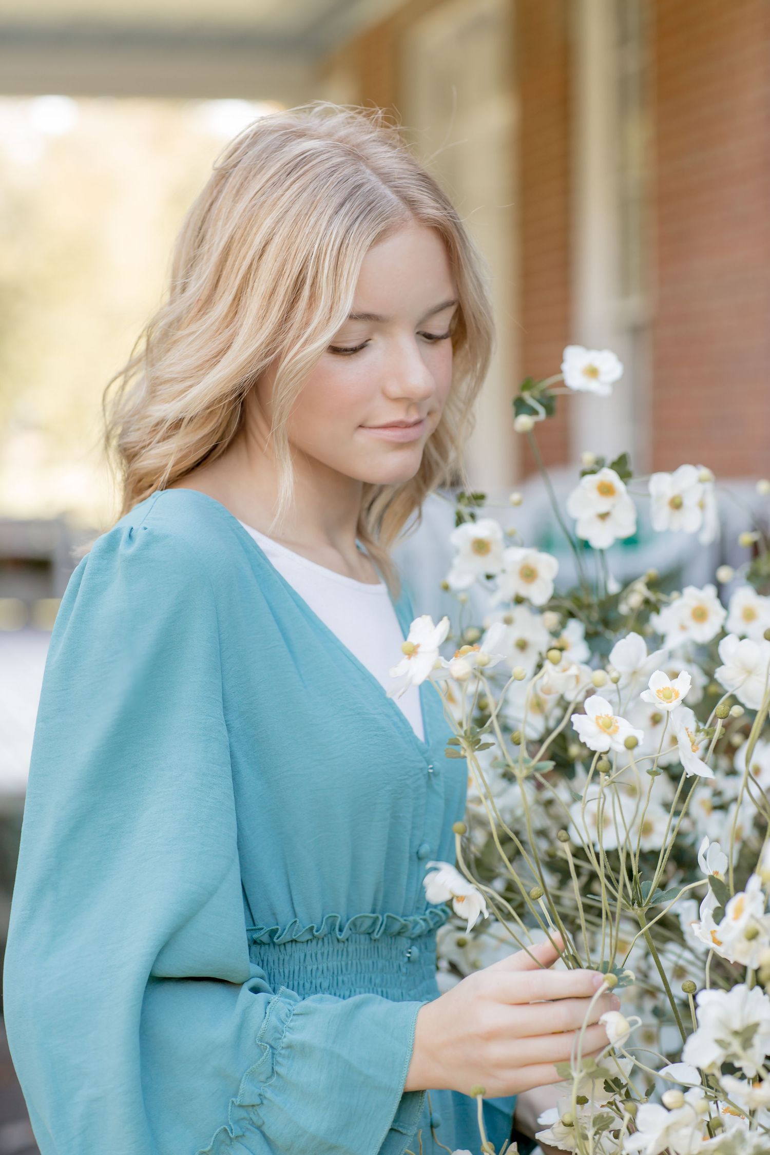 Young blonde woman in a teal dress gently holding white flowers outdoors in soft natural light.