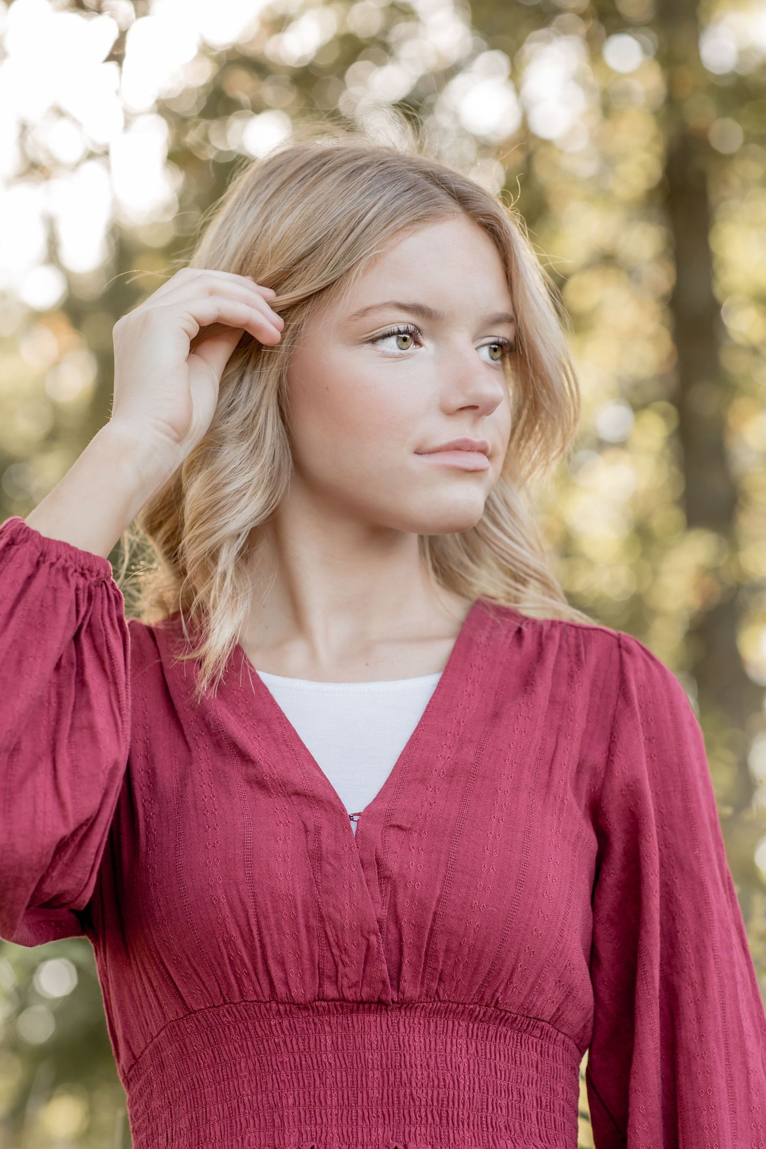 Blonde teenage girl in a burgundy dress tucking hair behind her ear with a thoughtful expression outdoors.