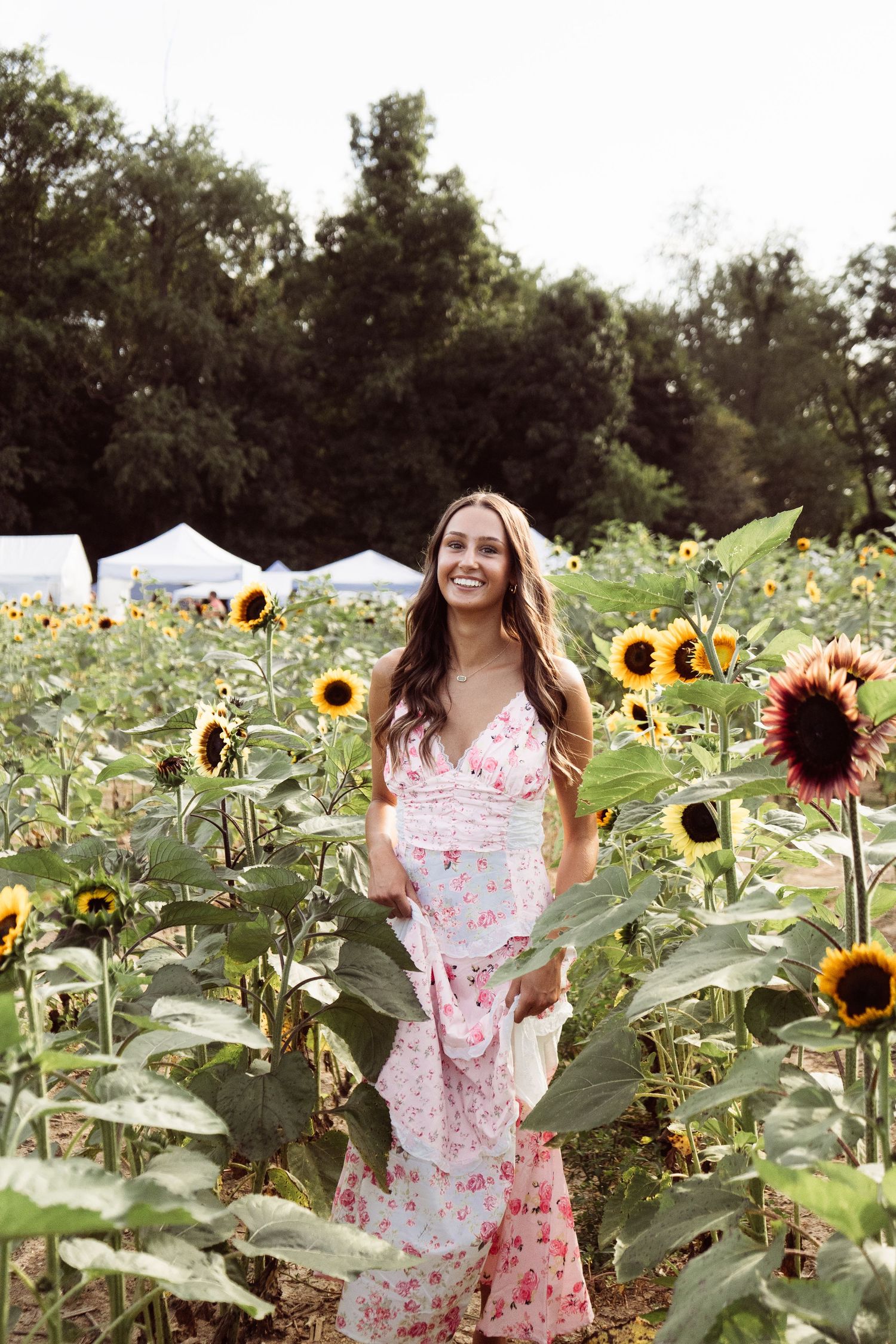 Smiling woman in pink floral maxi dress standing among vibrant sunflowers in field.