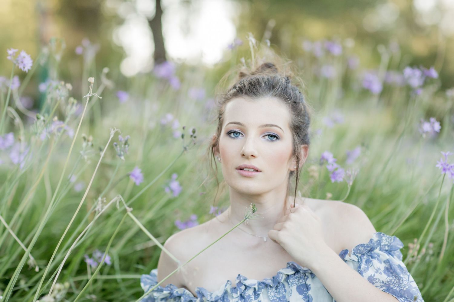 A young woman with blue eyes sits among purple wildflowers wearing an off-shoulder floral dress in a dreamy outdoor setting.