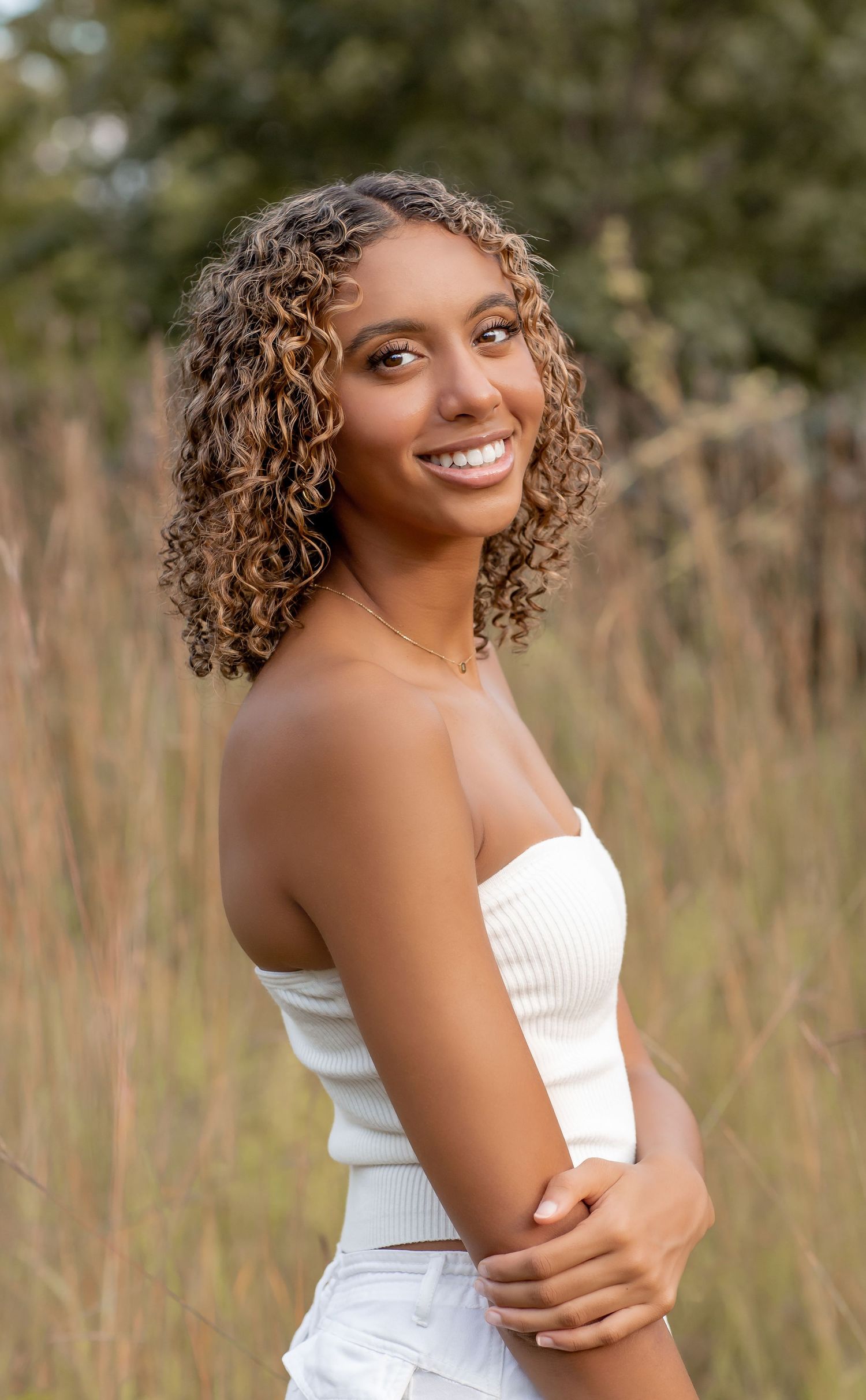 Young woman with curly highlighted hair smiling in a white strapless top in a golden grass field.