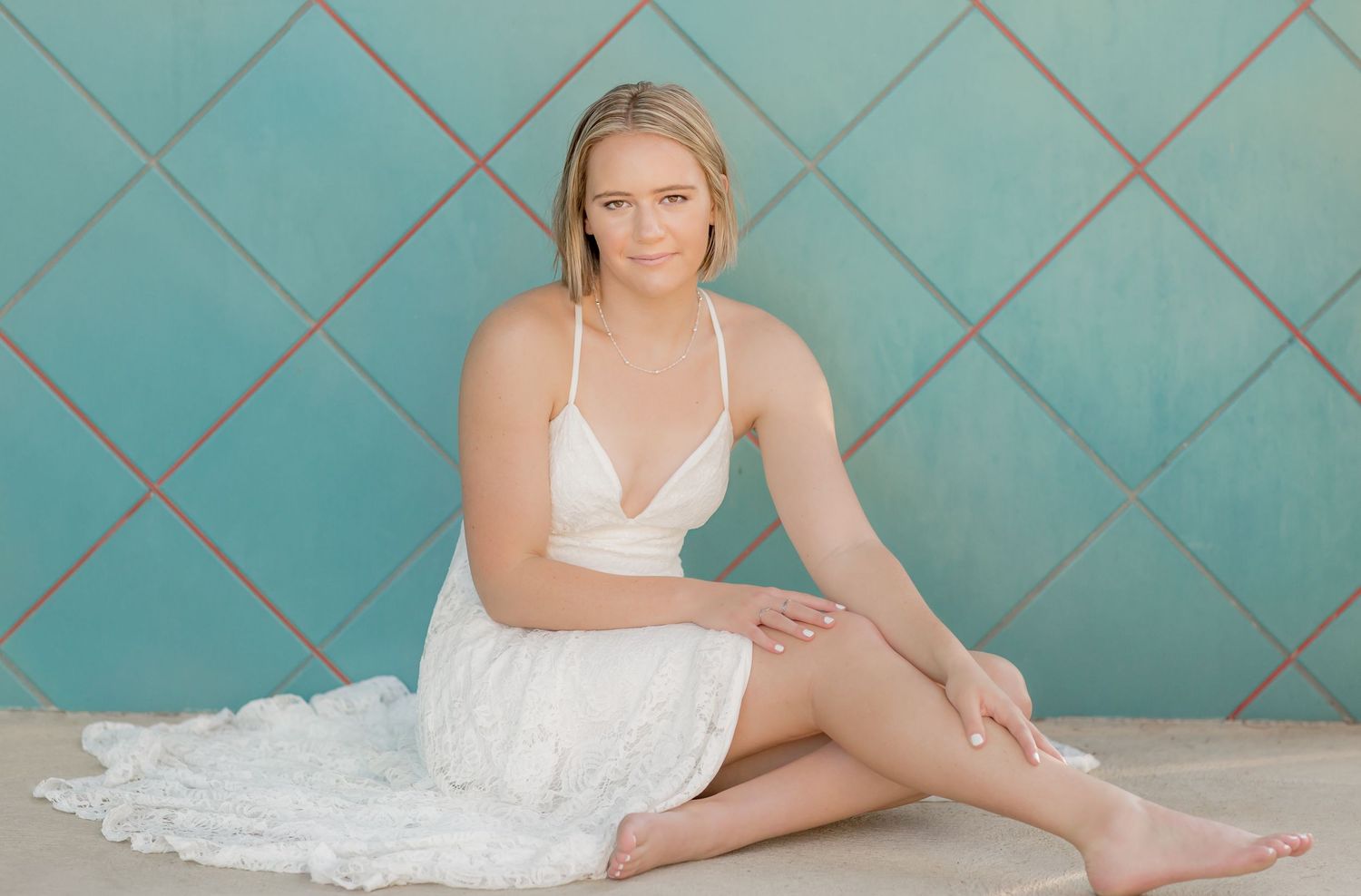 Blonde young woman in a white sundress sitting barefoot against a teal tiled wall indoors.