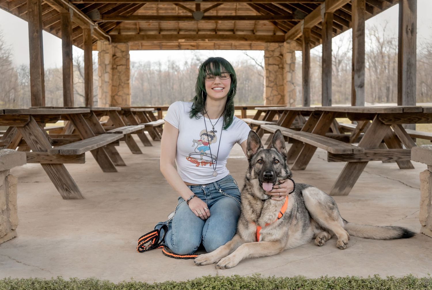 Smiling young woman with green hair sitting with a German Shepherd dog under a wooden pavilion.