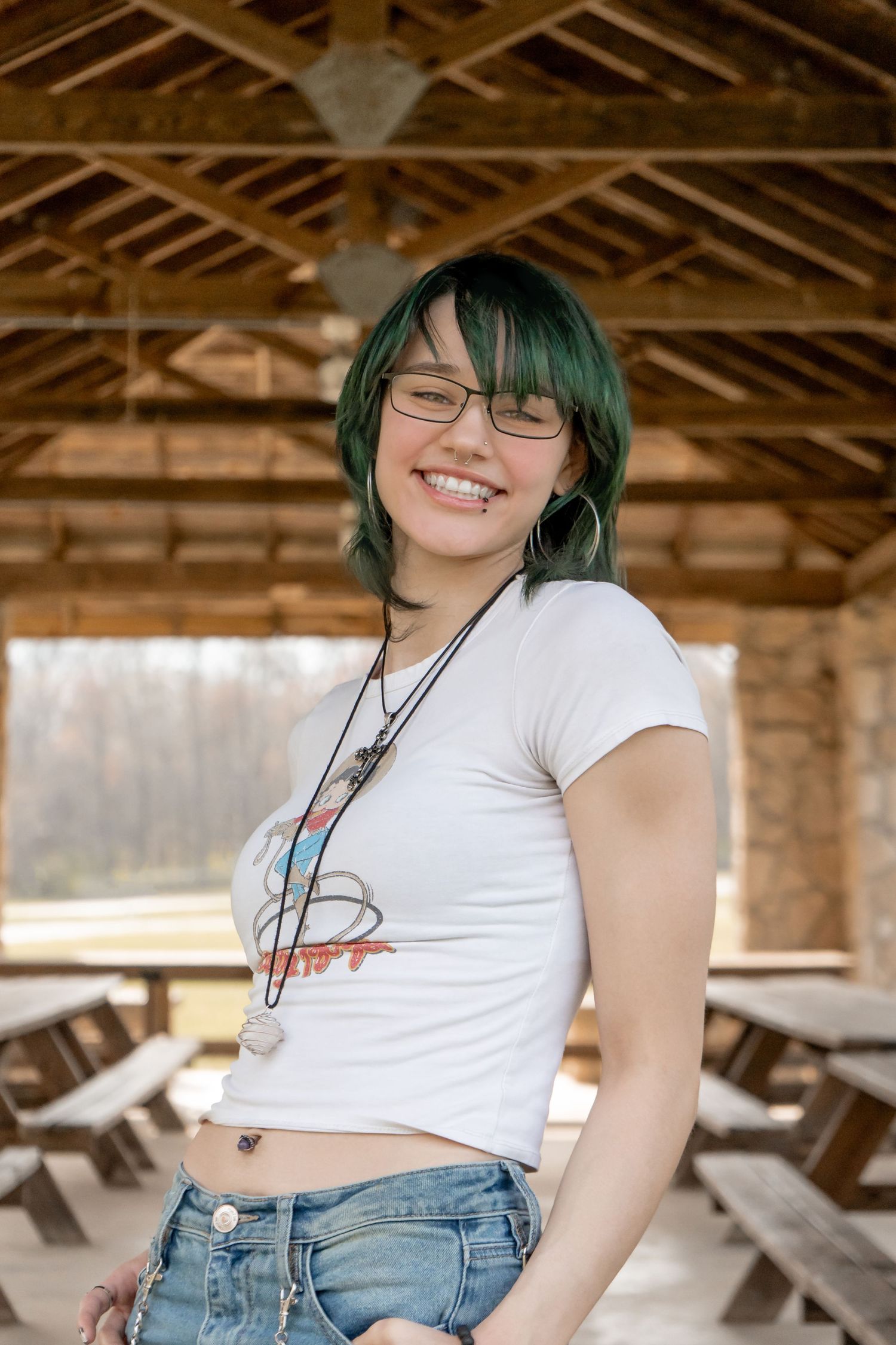 Smiling young woman with green hair and glasses wearing a white crop top and jeans in a pavilion.