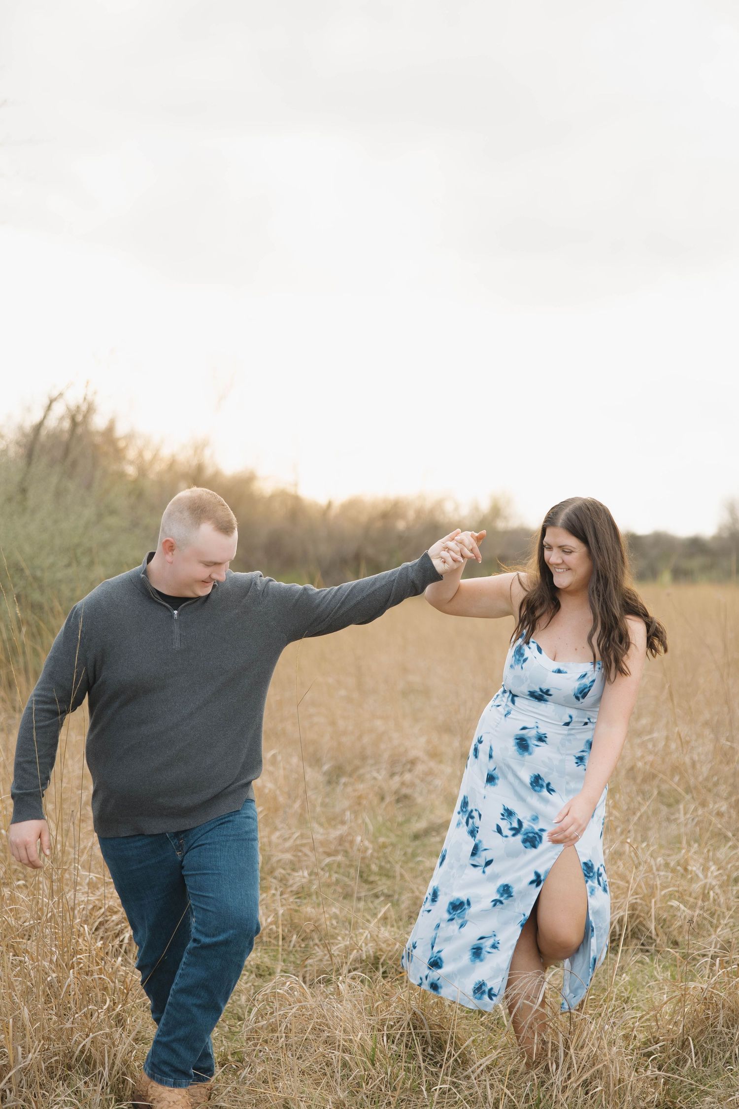 A couple walks hand in hand through a golden field at sunset, with her wearing a blue floral dress and him in casual attire.