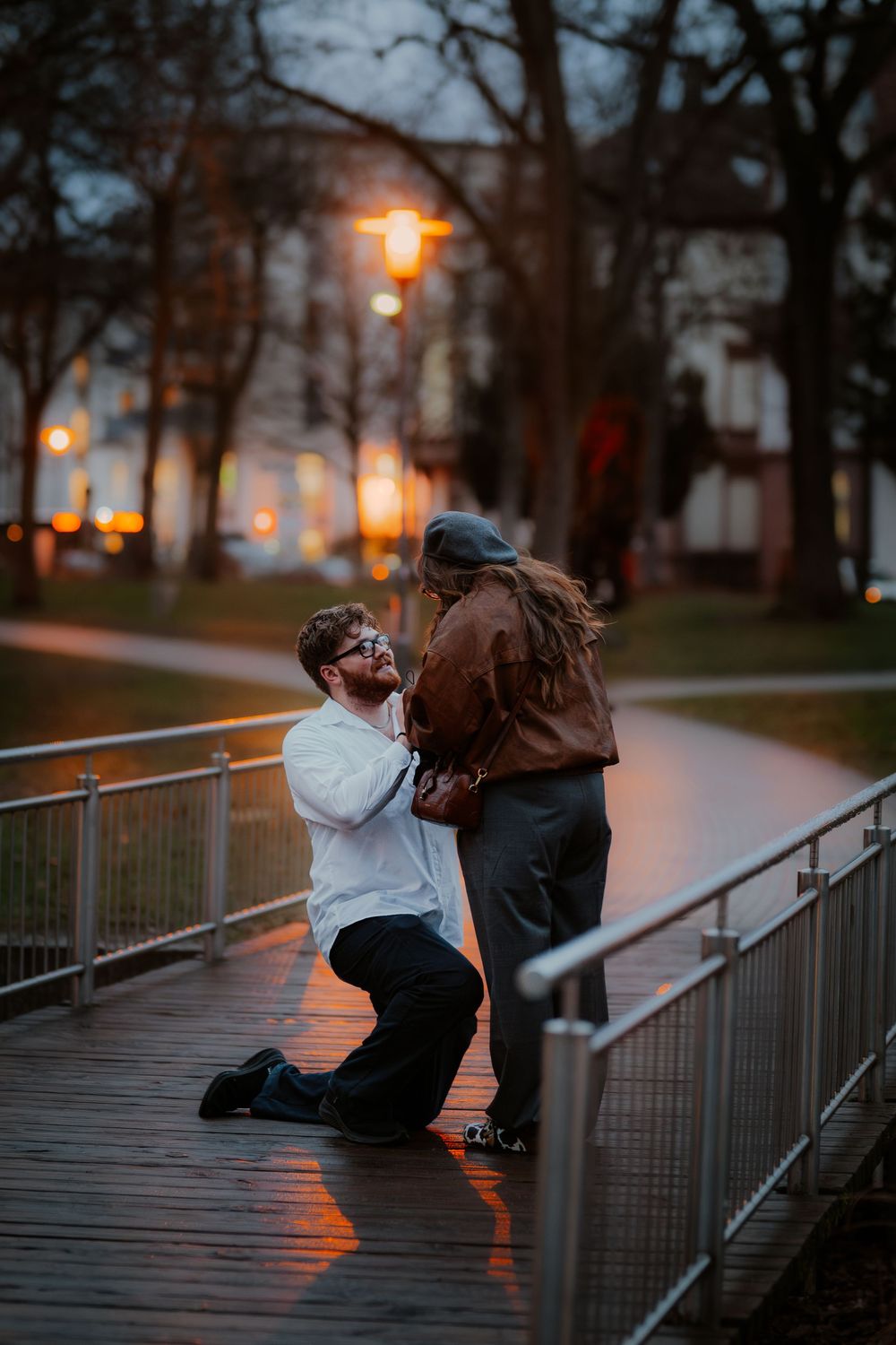 Couple sharing a romantic proposal moment on a wet bridge walkway at dusk with glowing street lights in the background.