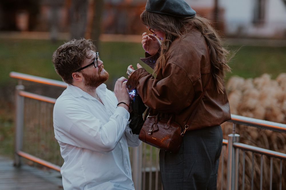 A couple shares a joyful moment during a proposal on a deck with fall foliage in the background.