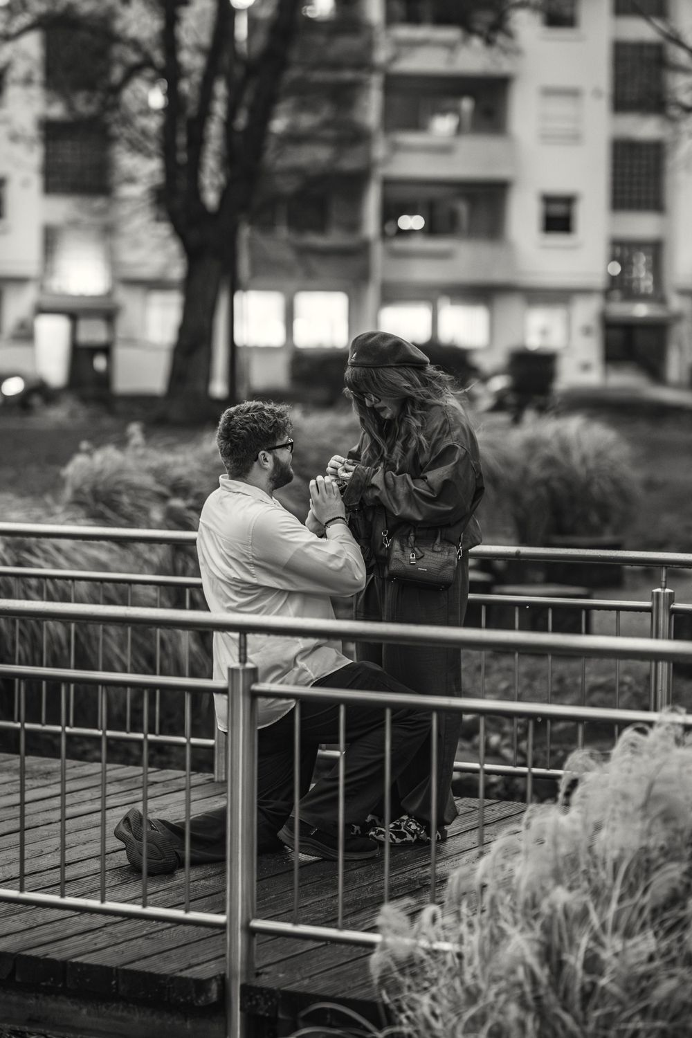 A romantic marriage proposal moment captured in black and white on a bridge deck with city buildings in the background.