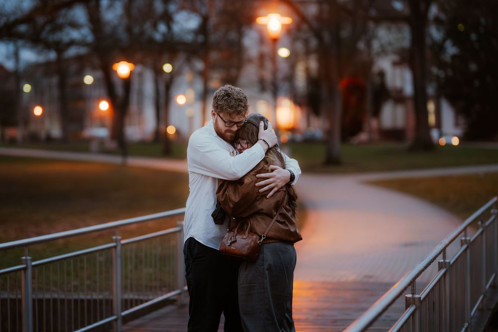 Two people embrace intimately on an illuminated walkway at dusk with glowing street lamps in the background.