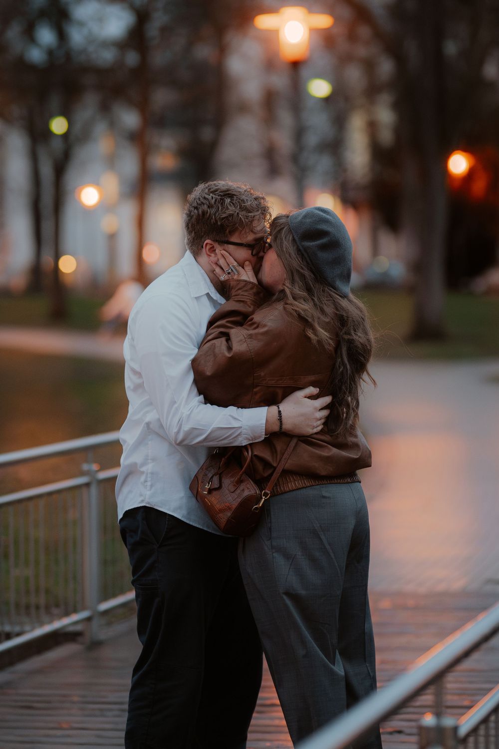 A romantic couple embrace on a bridge at dusk as street lamps glow warmly in the background.