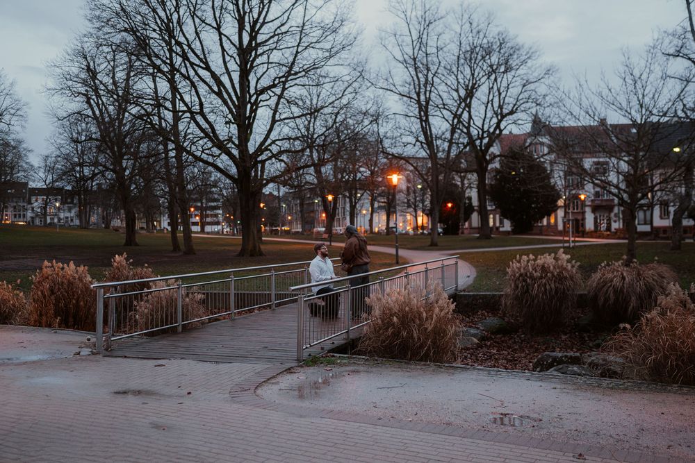 A person stands on a pedestrian bridge in a park at dusk with bare trees and city buildings in the background.