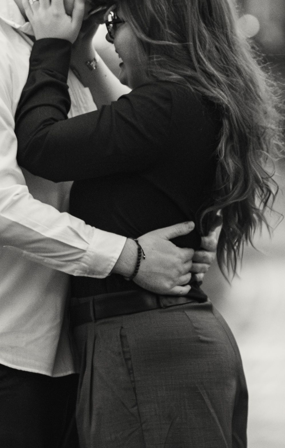 Black and white photo of a romantic embrace from behind with man's hand on woman's waist as she touches her hair.
