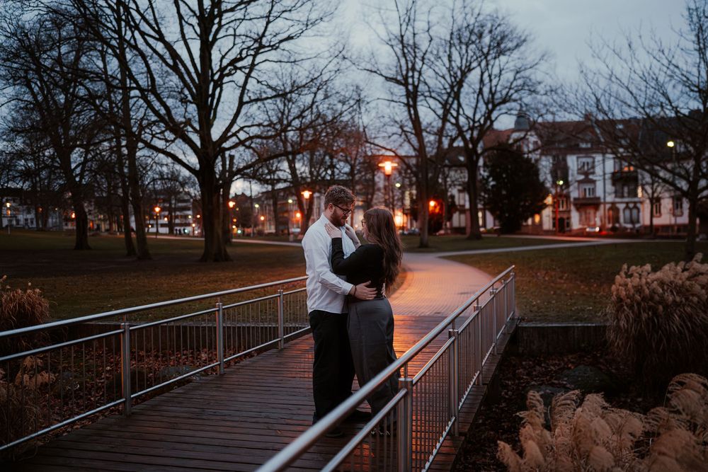 Couple embraces on wooden bridge at dusk with illuminated buildings glowing in background during romantic evening.