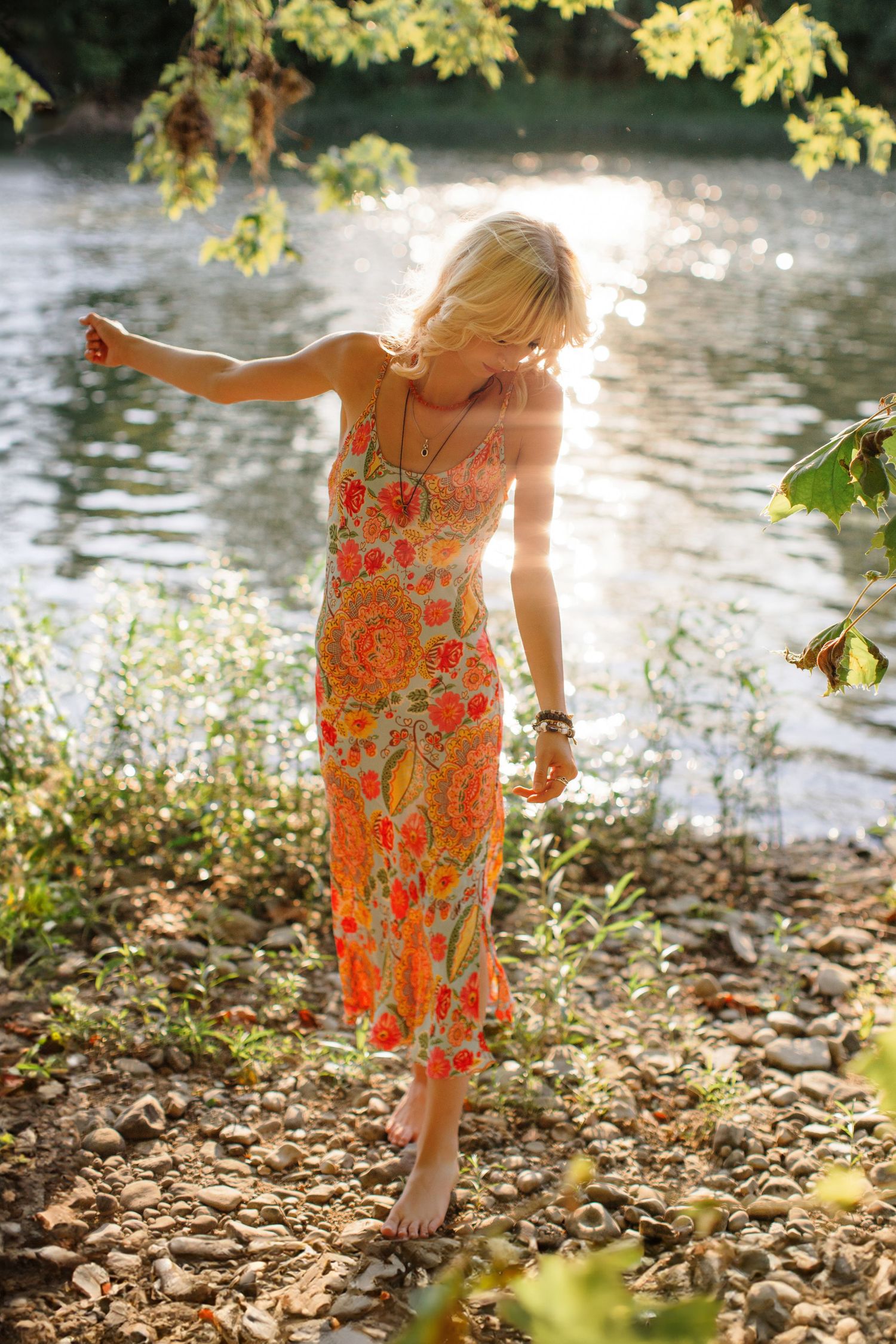 Dancing figure in a coral floral maxi dress captured in sunlit moments by the riverbank.