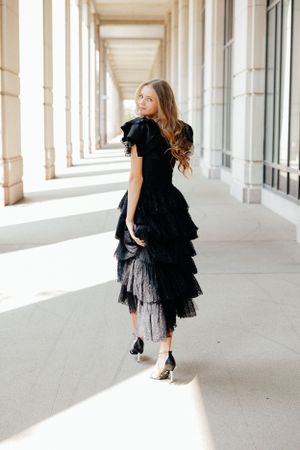 A series of dynamic poses in a black ruffled dress along a bright columned corridor.