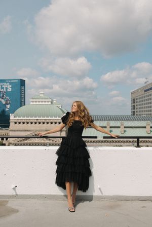 Woman in black dress poses dramatically on rooftop with urban skyline backdrop.
